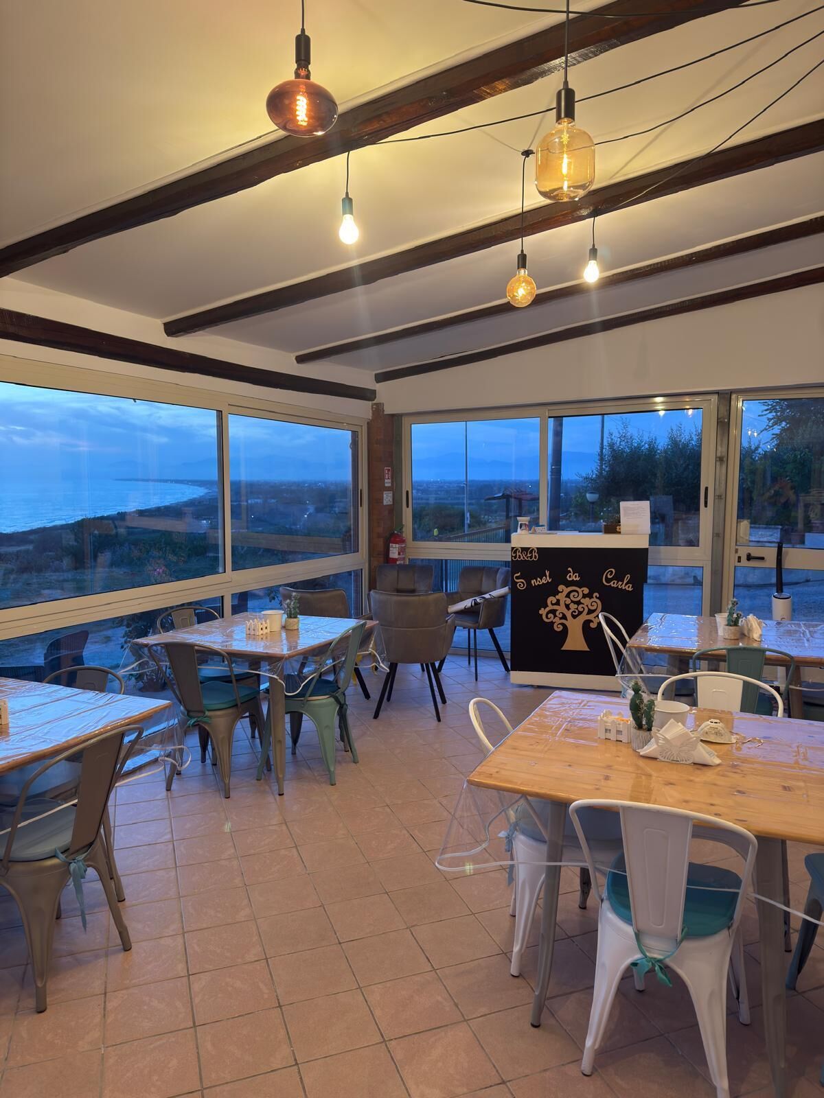 Comedor con mesas y sillas, ventanas amplias con vistas y lámparas colgantes.