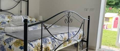 Une chambre à coucher avec un lit en métal, une literie à motifs floraux et une vue sur un paysage verdoyant, visible à travers une porte vitrée.