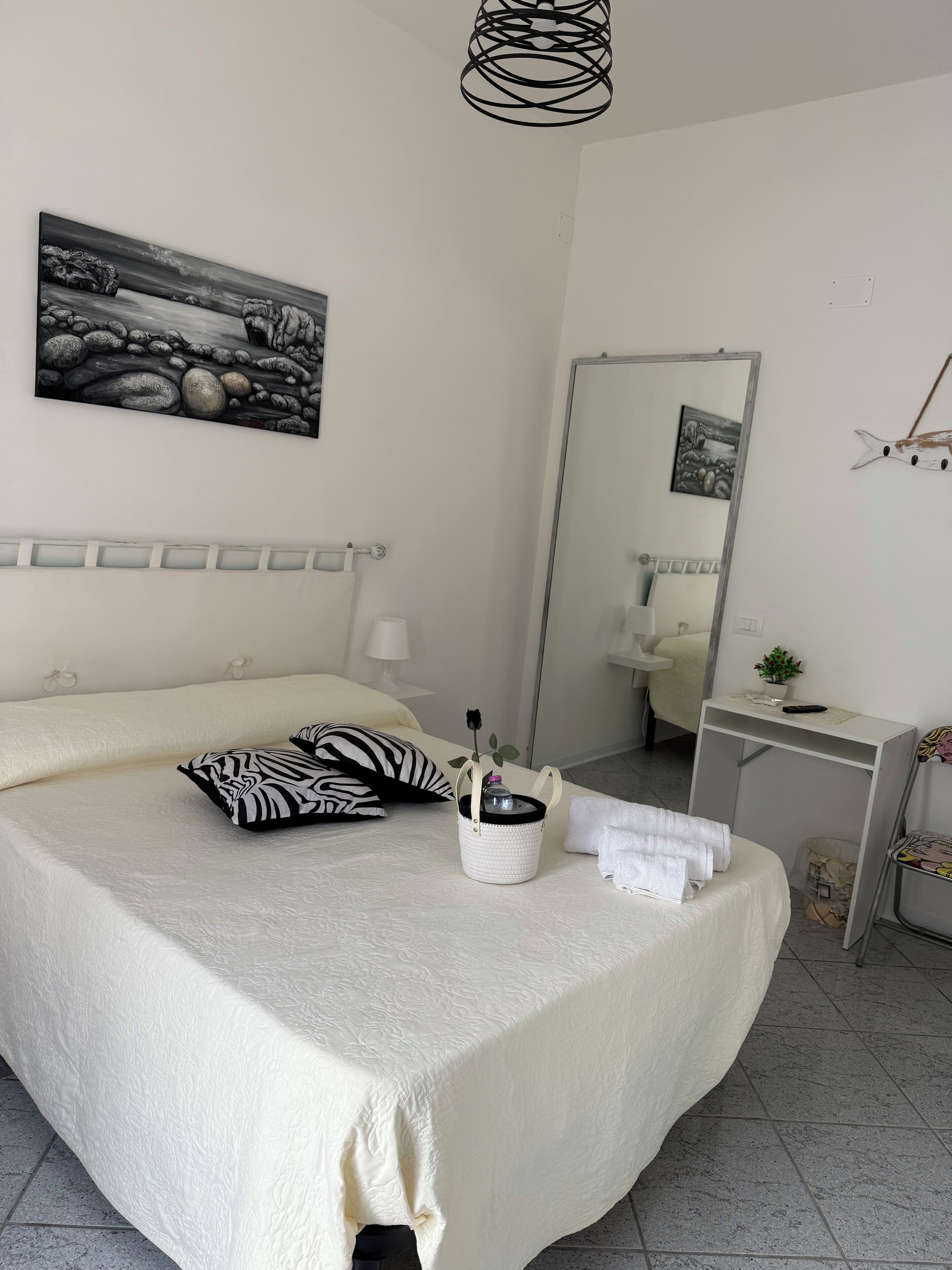 A neatly made bed with a zebra-patterned pillow, a small white basket with a rose, and a white towel. A large mirror and a framed picture are on the wall.
