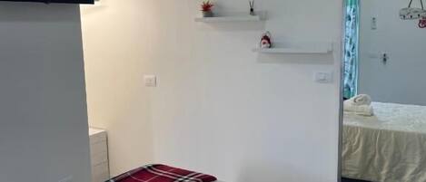 A hotel room with a bed, a red plaid blanket, a white pillow, and a small shelf with decorative items.