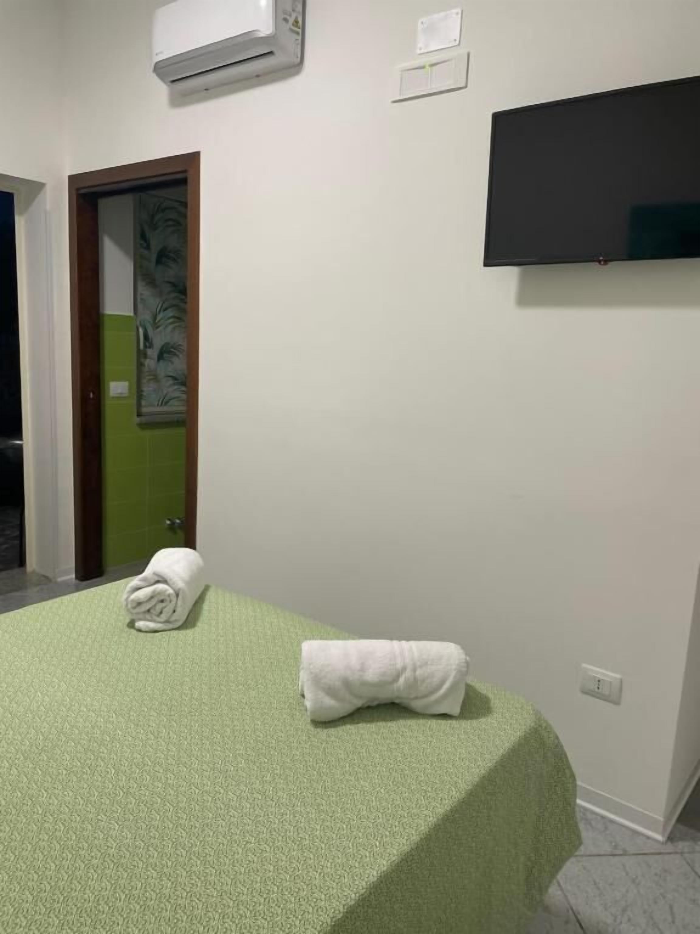 A room with a bed covered in a green blanket, a flat-screen TV mounted on the wall, and an air conditioning unit above the bed.