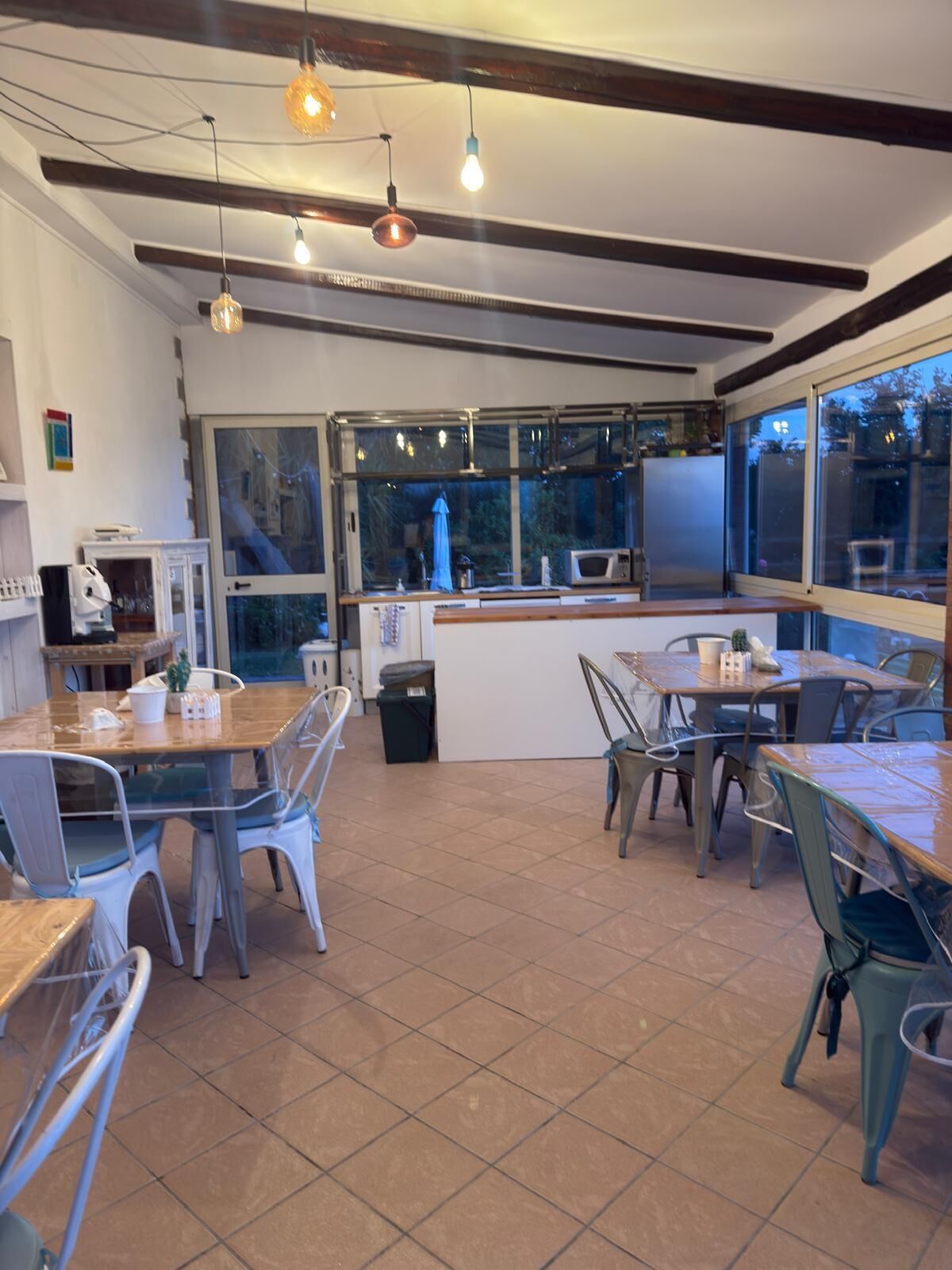 A restaurant with a kitchen area, dining tables, chairs, and hanging lights.