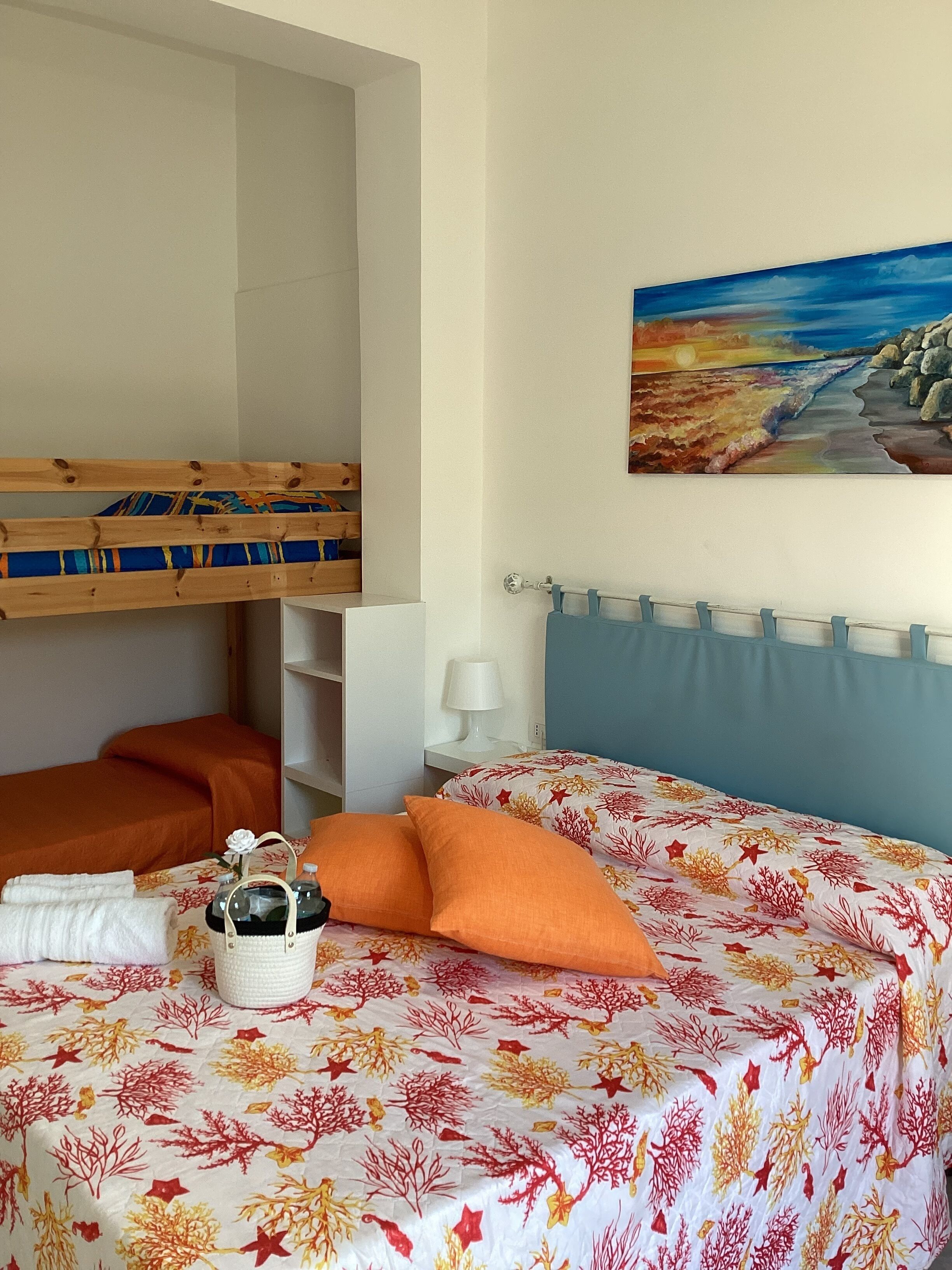 A bedroom with a bunk bed, a bed with a floral bedspread, a bedside table with a lamp, and a painting on the wall.