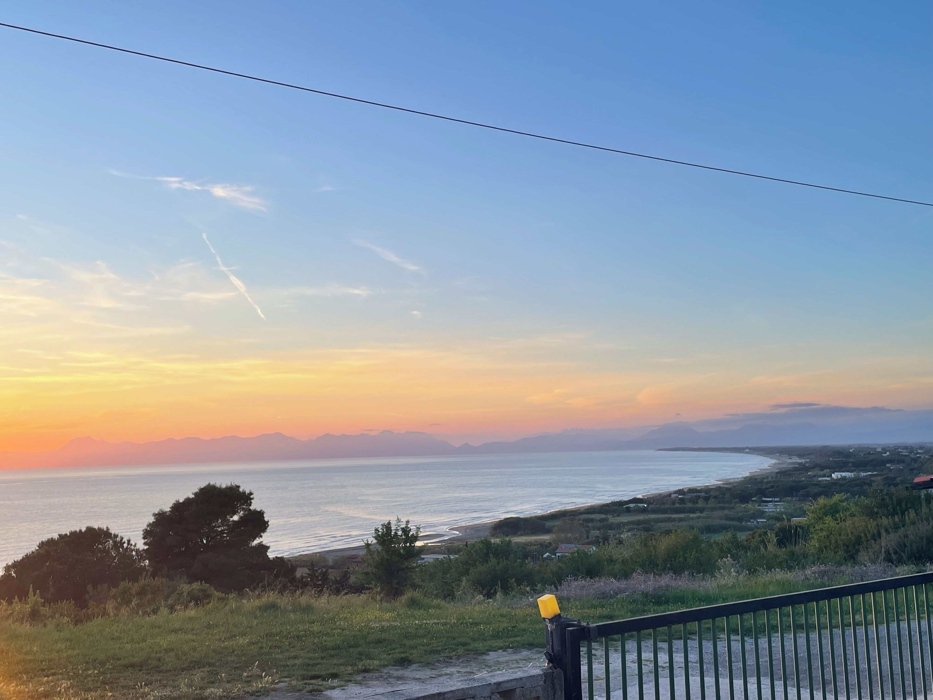 A coastal landscape with a clear sky, a body of water, and a grassy area with a fence.