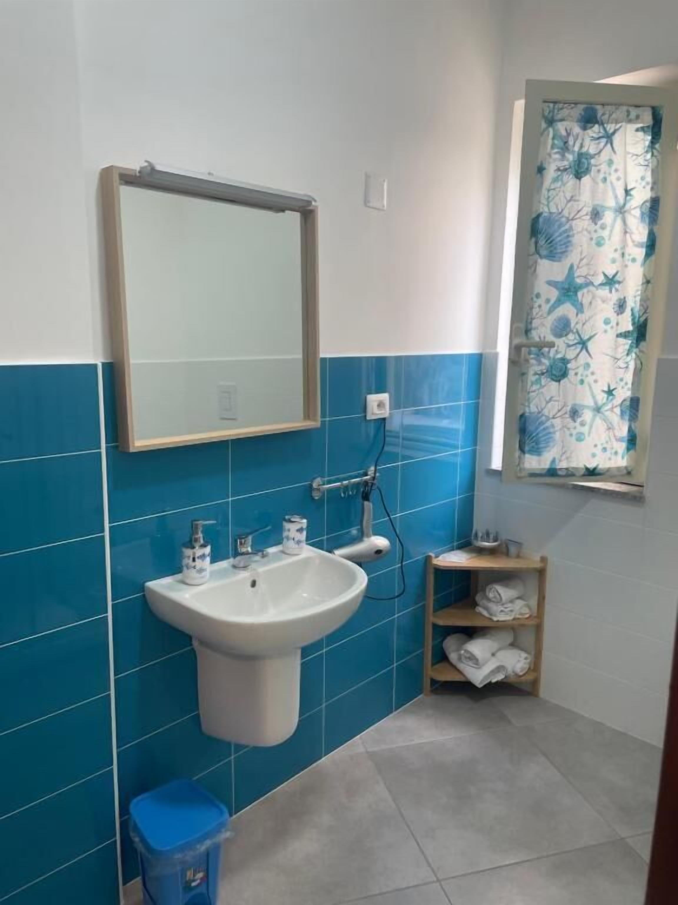 A bathroom with a white sink, blue tiles, a mirror, a towel rack, and a window with a floral curtain.