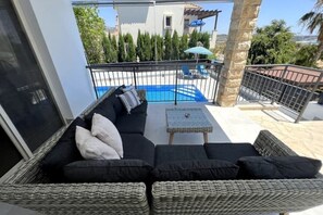 Ein Balkon mit einer Rattan-Couch, einem kleinen Tisch und einem Pool im Hintergrund.