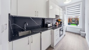 A modern kitchen with white cabinets, a black countertop, and a window with blinds. - Bright & Stylish Flat in the Heart of Islington (London)