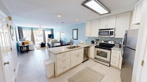 A modern kitchen with white cabinets, a stainless steel refrigerator, a microwave, and an oven. There is a dining area with a table and chairs, and a living room with a sofa and a TV. - Maravilla 1213 - Dog Friendly and Beach Service! (Miramar Beach)