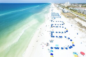 Une plage de sable blanc bordée d’une eau d’un bleu cristallin. De nombreux parasols bleus et des chaises de plage sont alignés le long du rivage.