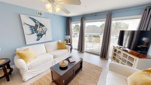 A living room with a white sofa, a coffee table, a TV, and a balcony with curtains. - Maravilla 1213 - Dog Friendly and Beach Service! (Miramar Beach)