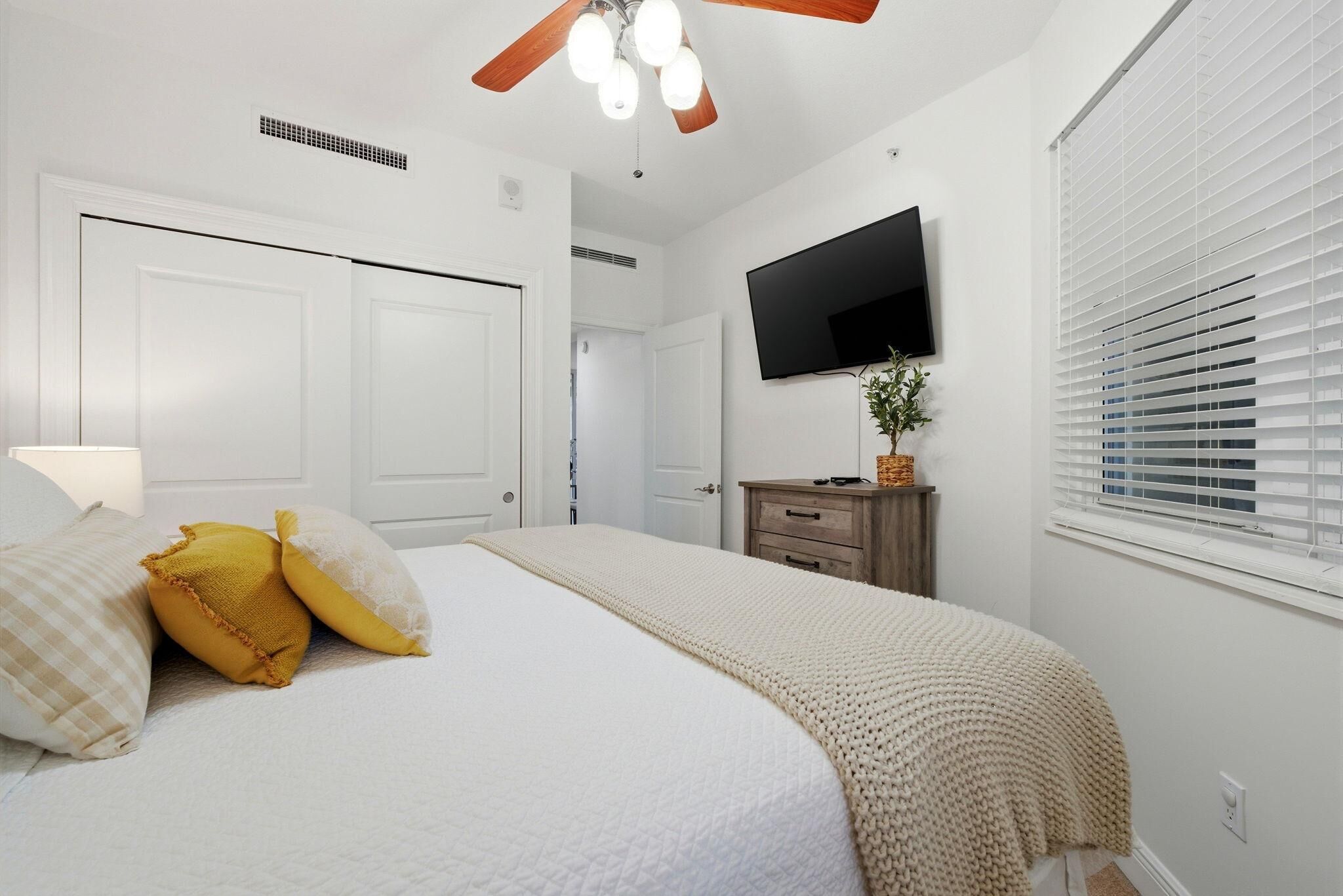Un dormitorio con cama, ventilador de techo, televisor y ventana con persianas.