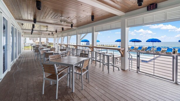 A covered outdoor area with wooden flooring, tables, and chairs, overlooking a beach with blue umbrellas. - Lost Key B203 (Pensacola)