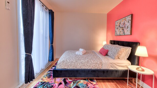 Ein Schlafzimmer mit einem Bett, einem Nachttisch, einer Lampe, einem farbenfrohen Teppich und einem Fenster mit Vorhängen.