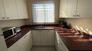 Eine kleine Küche mit weißen Schränken, einer dunklen Arbeitsplatte, einem Fenster mit Jalousien und einer Mikrowelle.