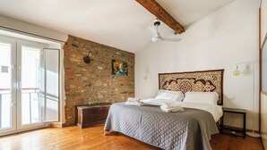Un dormitorio con pared de piedra, una cama con colcha acolchada, un ventilador de techo y una mesita de noche con lámpara.