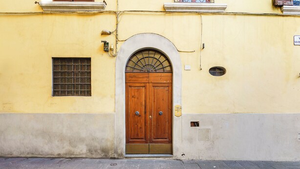 Un edificio amarillo con una puerta doble de madera, una pequeña ventana con celosía metálica y una acera de baldosas.