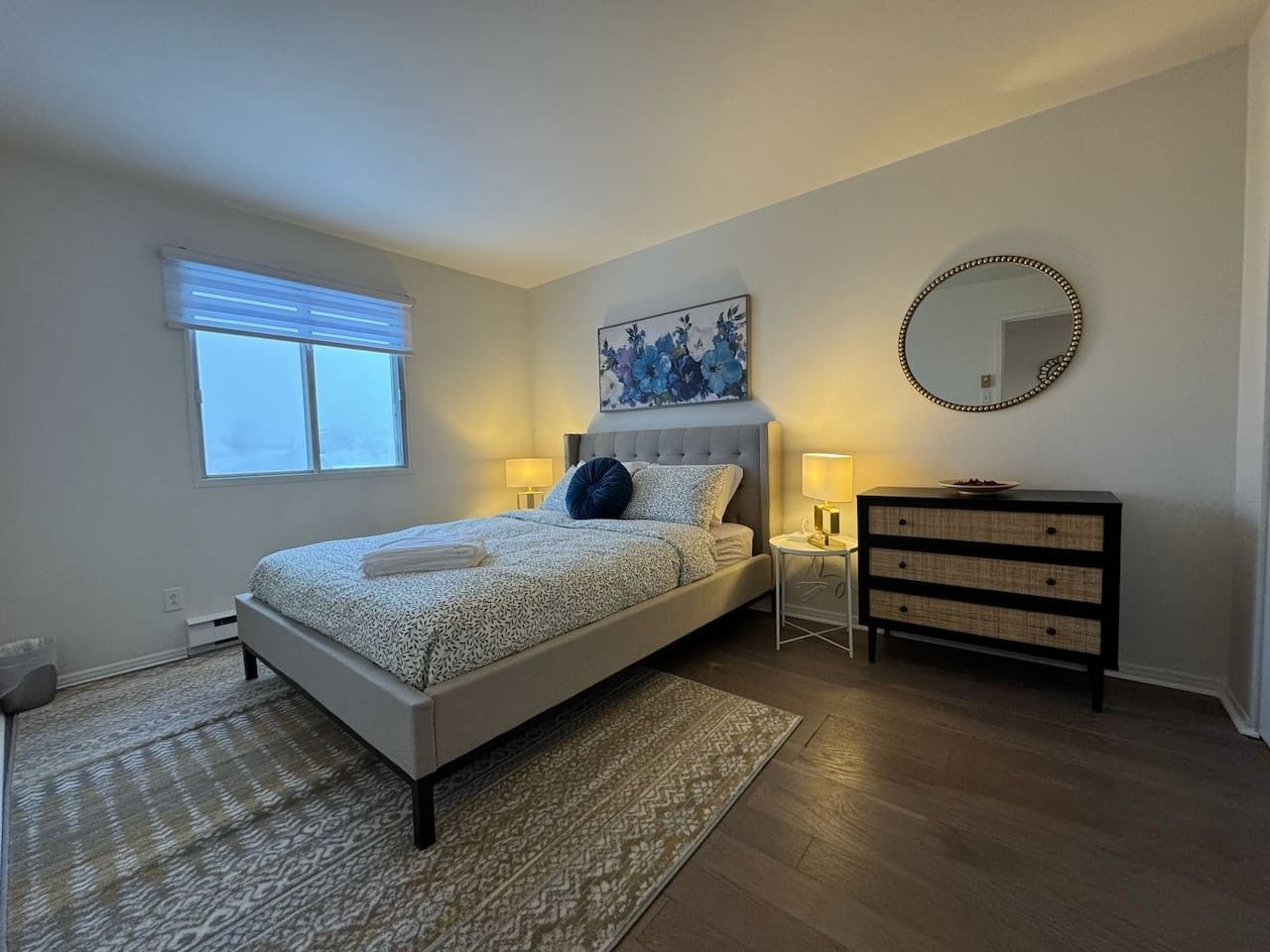 Een slaapkamer met een bed, nachtlampjes, een dressoir, een spiegel en een raam met jaloezieën.