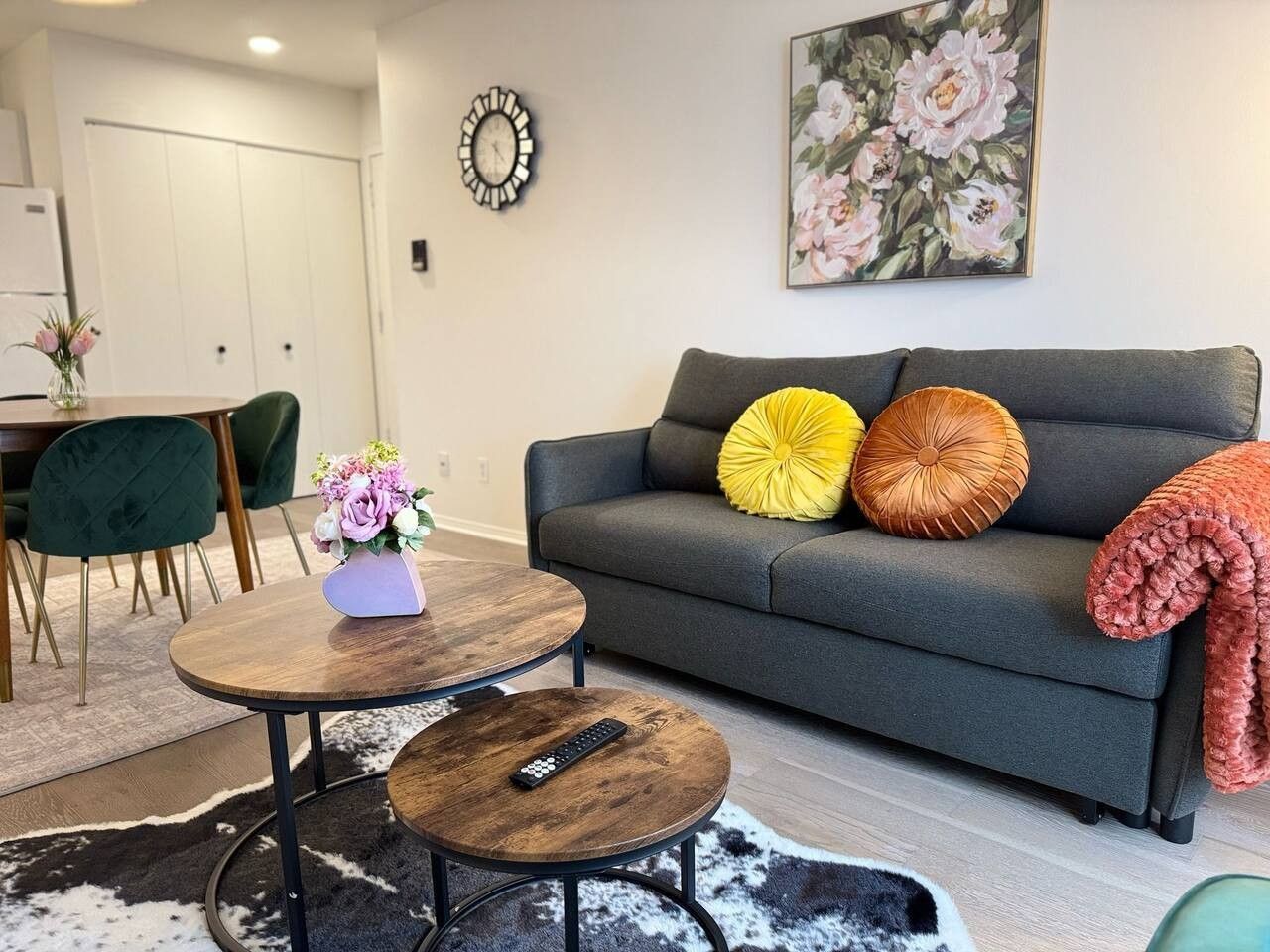 Een woonkamer met een grijze bank, twee ronde houten tafels, een afstandsbediening en een bloemrijk schilderij aan de muur.