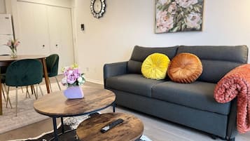 Een woonkamer met een grijze bank, twee ronde houten tafels, een afstandsbediening en een bloemrijk schilderij aan de muur.