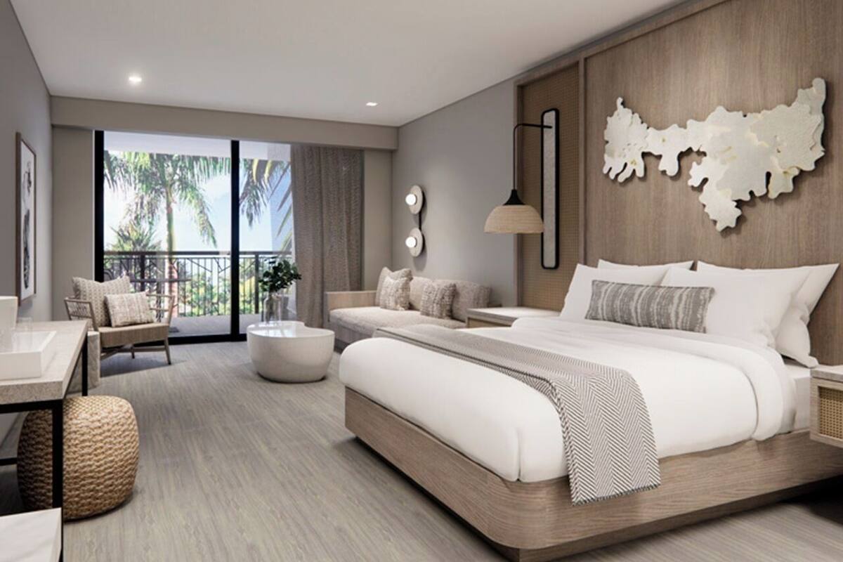 Una habitación de hotel moderna con una cama grande, una zona de estar con sofá y sillones, una mesa de comedor y un balcón con vistas a palmeras.