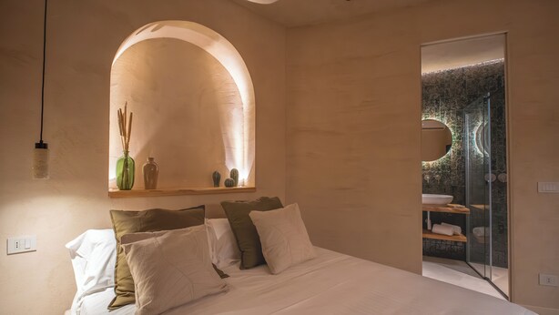 A bedroom with a large bed, a wooden shelf with decorative items, a mirror, and a glass door leading to a bathroom. - Essenza Boutique Apartments (Cefalù)