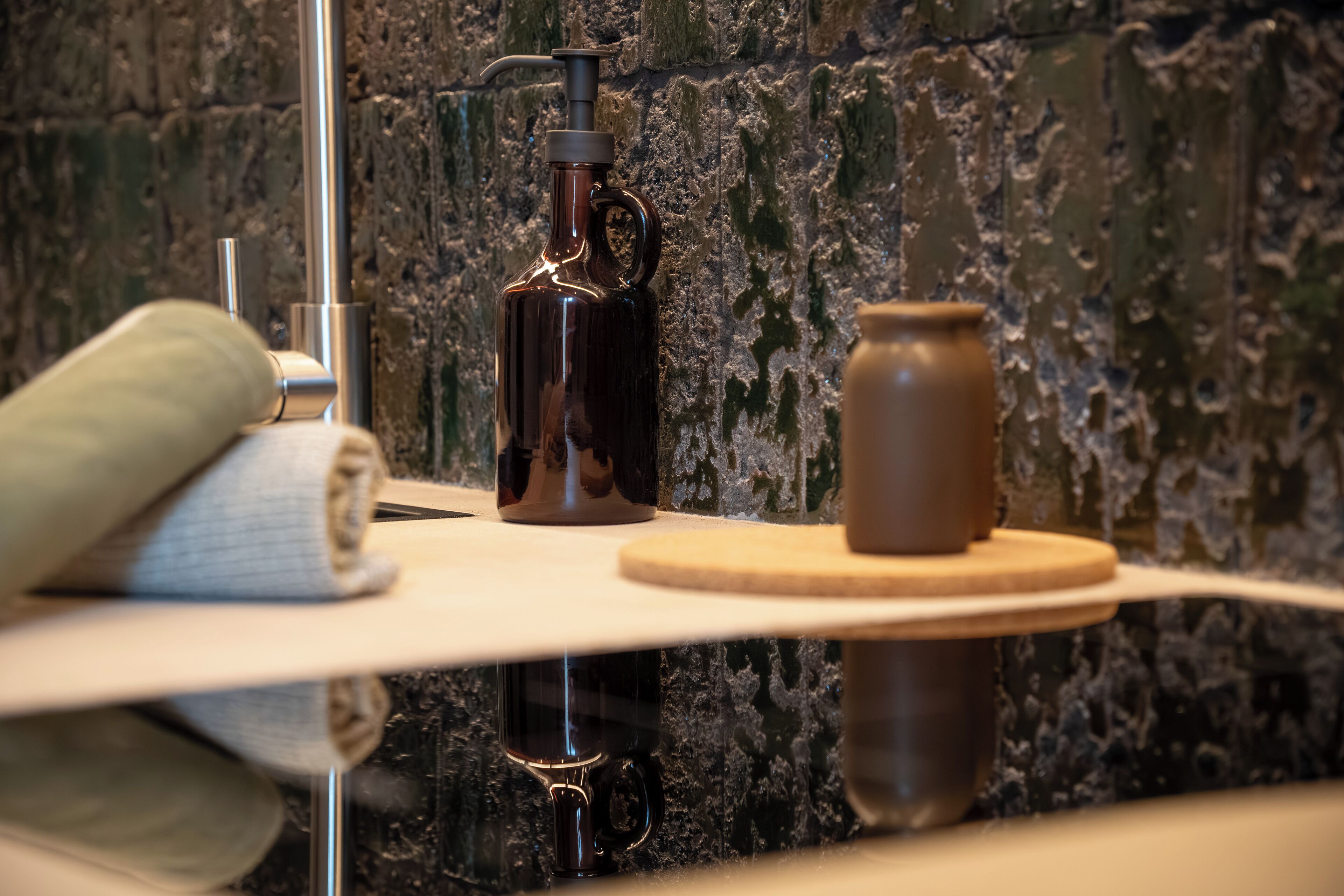 A bathroom with a marble wall, a towel, a soap dispenser, and a small jar on a wooden shelf.