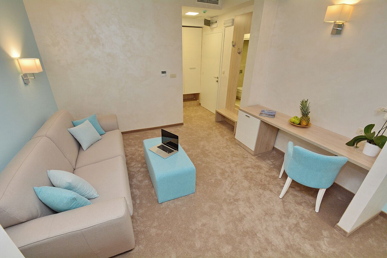 A compact living space with a beige sofa, a small blue ottoman with a laptop, a light blue chair, and a desk with a plant and fruit.