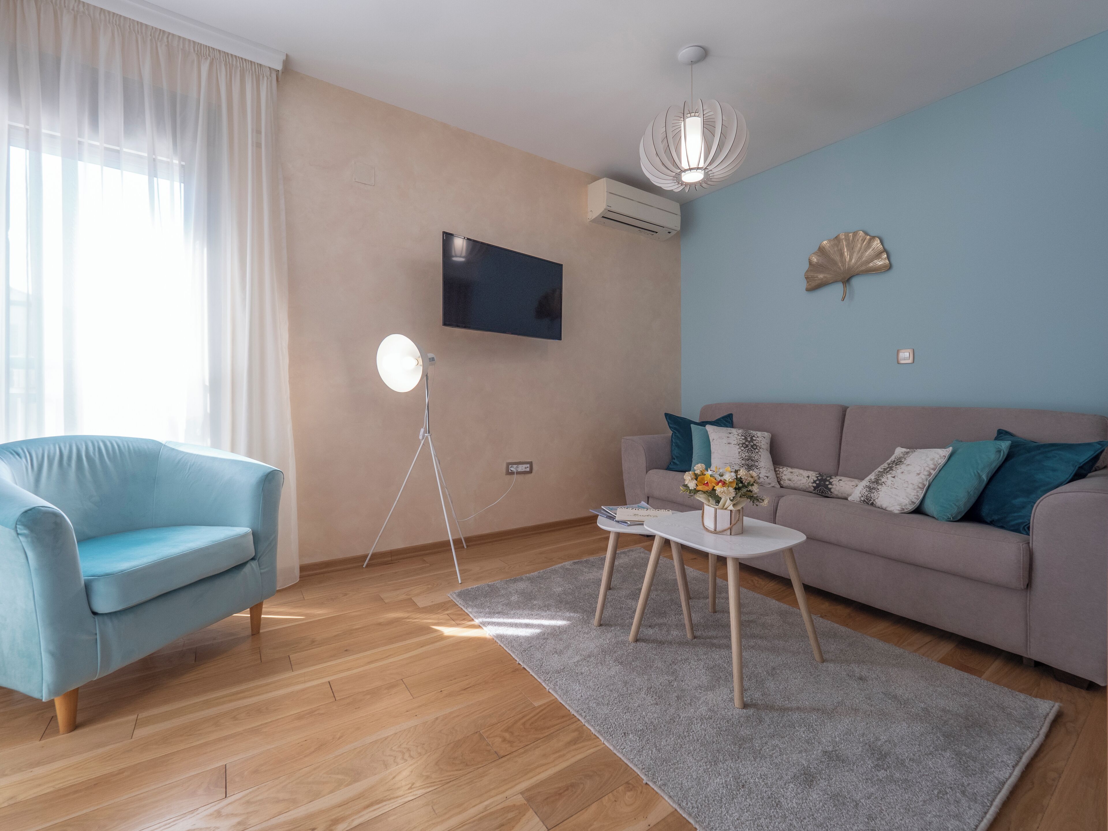 A modern living room with a blue sofa, a light blue armchair, a white coffee table with a vase of flowers, a floor lamp, and a mounted TV.