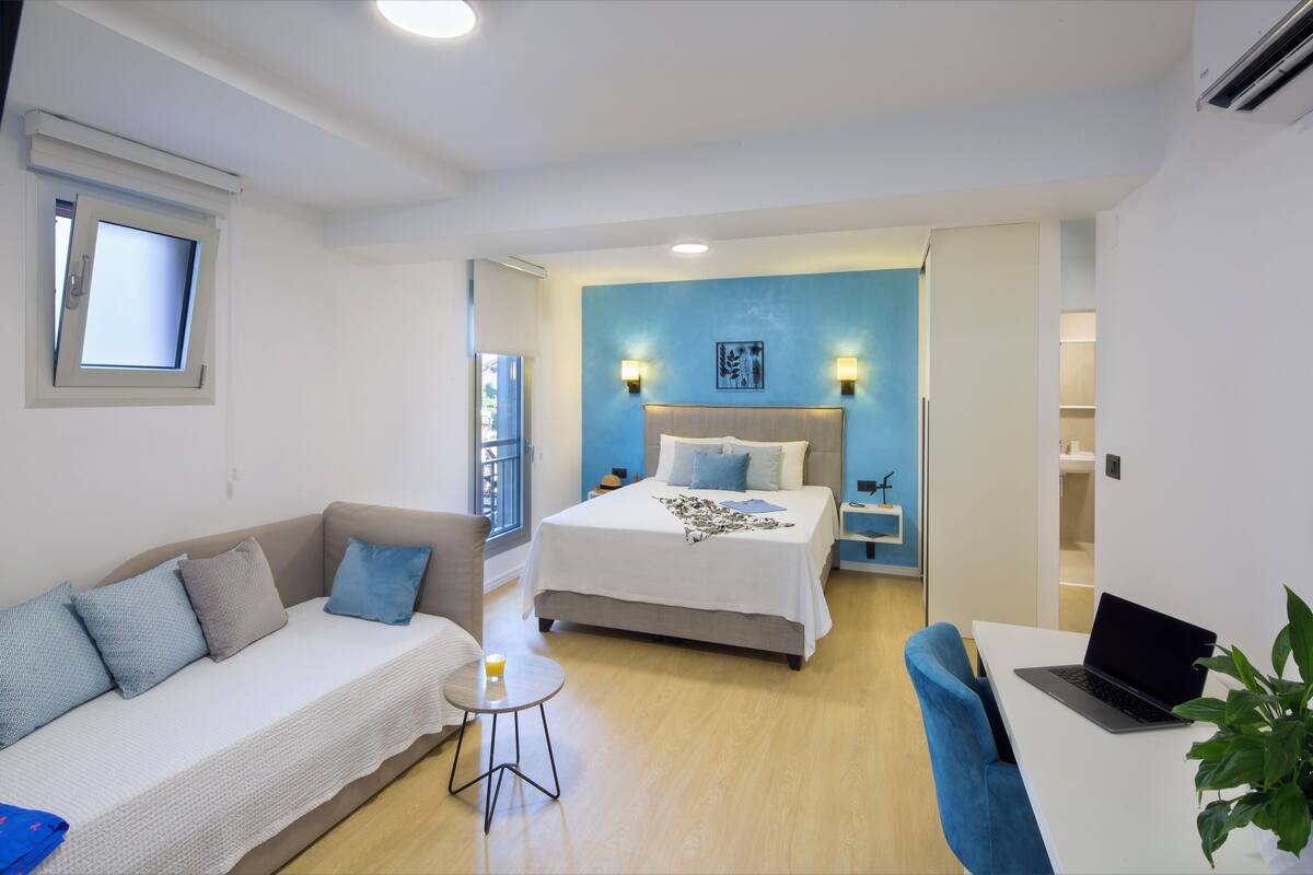 A modern hotel room with a bed, a sofa, a small table, and a laptop on a desk.