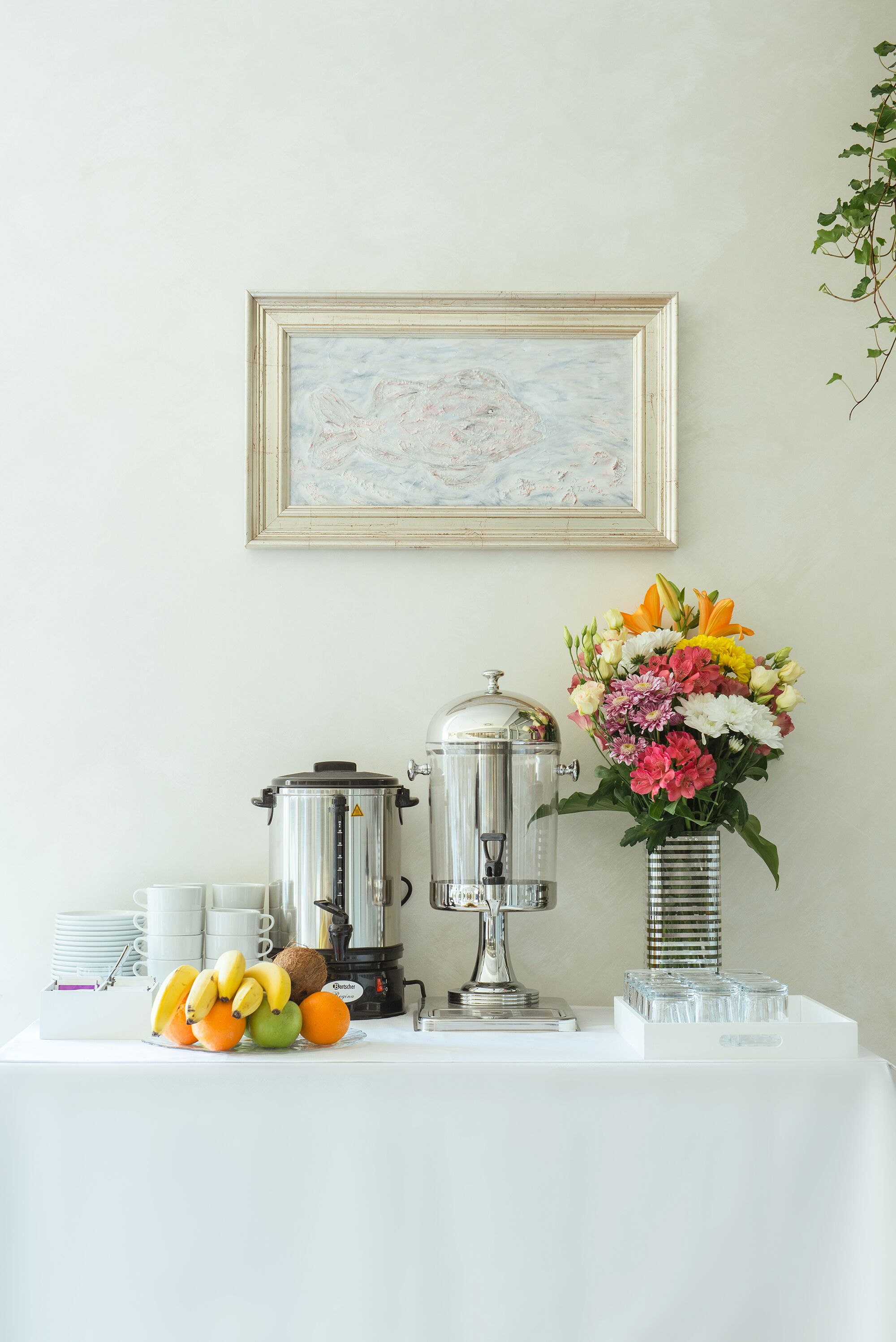 A breakfast setup with a coffee maker, a fruit bowl, and a vase of flowers on a white table.
