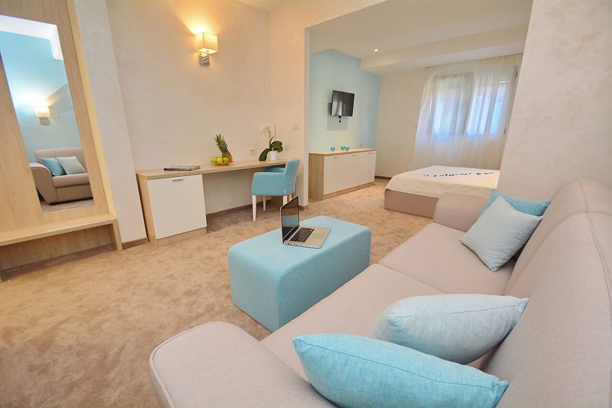A modern hotel room with a sofa, a desk with a laptop, a bed, and a TV mounted on the wall.