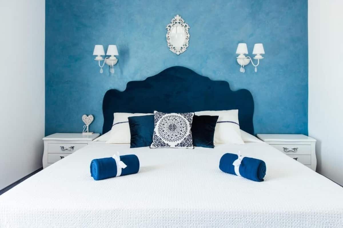A neatly made bed with a blue headboard, white bedding, and decorative pillows. There are two bedside tables with lamps and a heart-shaped decoration on one.