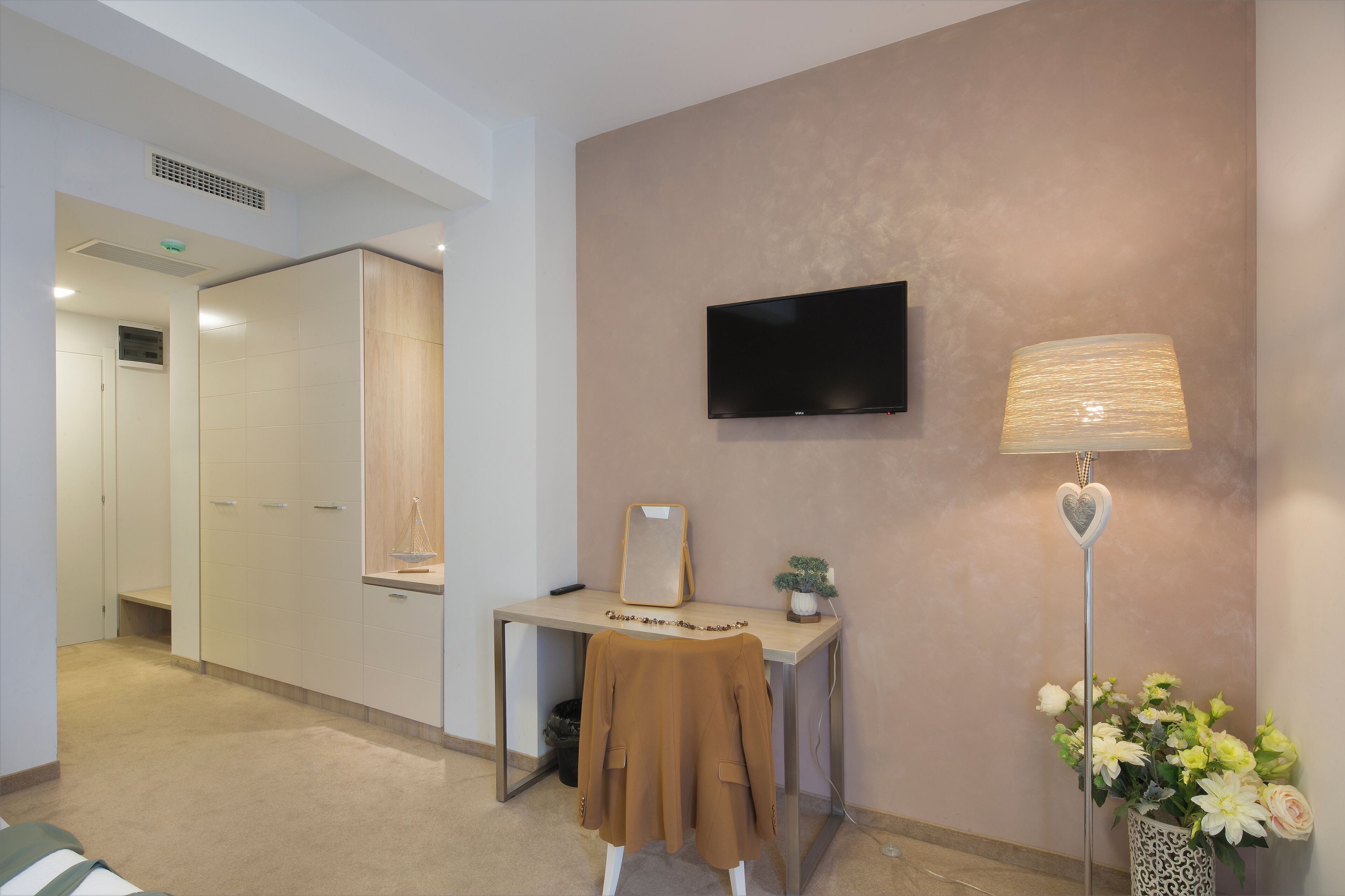 A hotel room with a desk, a flat-screen TV, a lamp, and a flower arrangement.