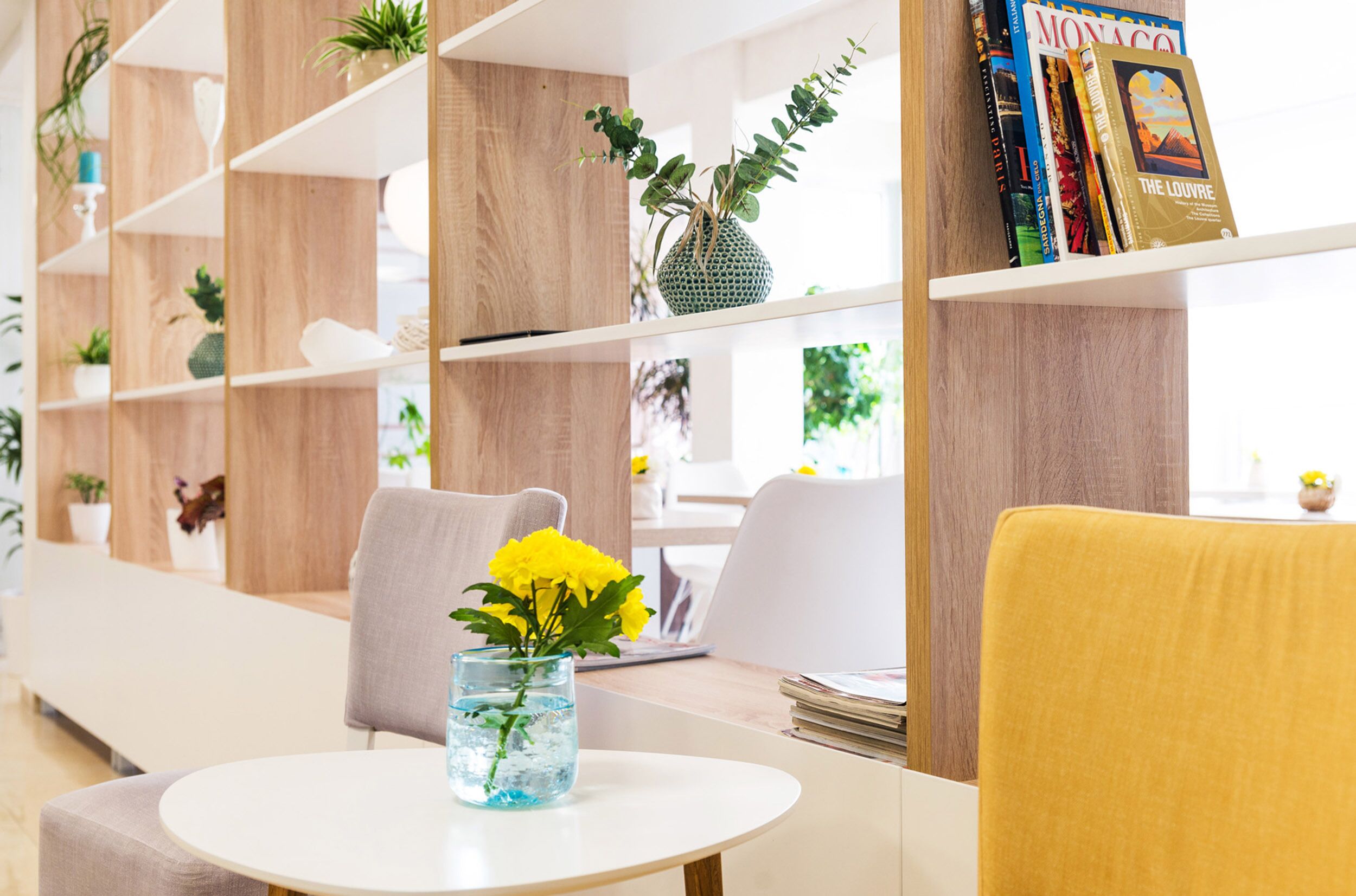 A modern interior with a round table, a vase with yellow flowers, and a bookshelf with books and decorative items.