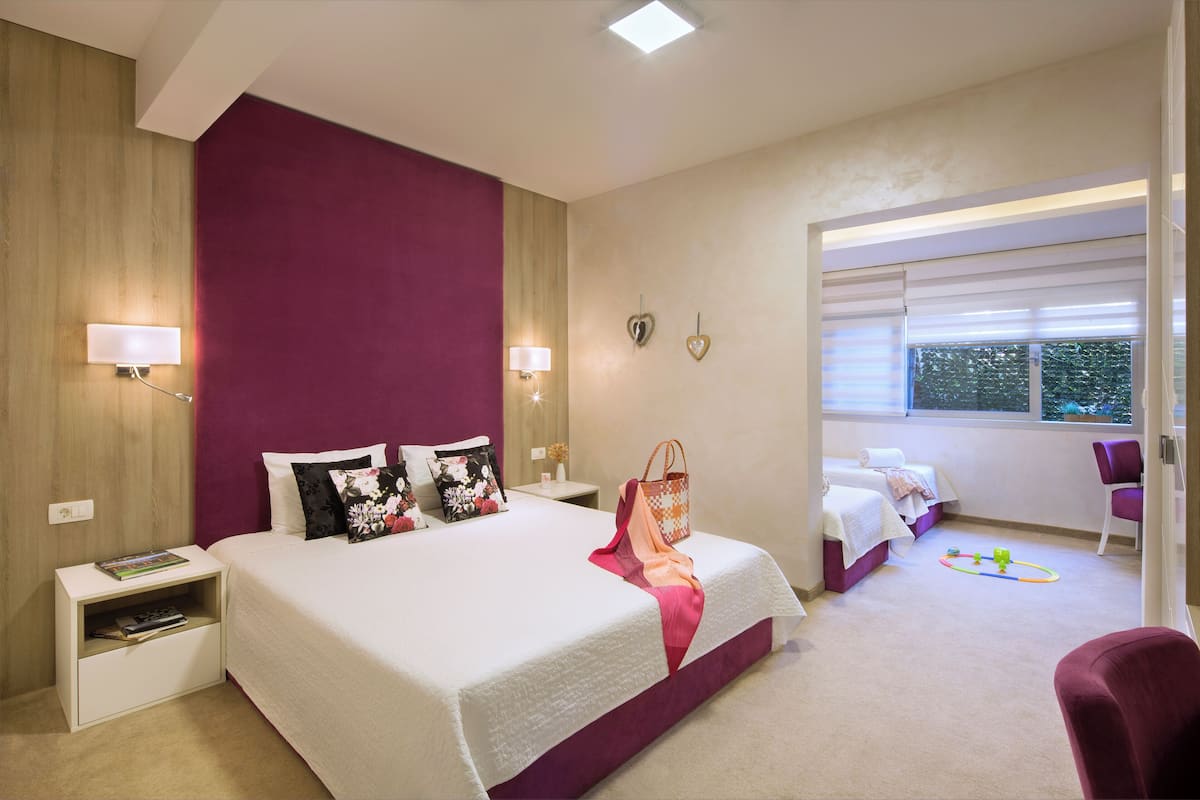 A modern bedroom with a large bed, bedside tables, a nightstand, a chair, and a window with blinds.