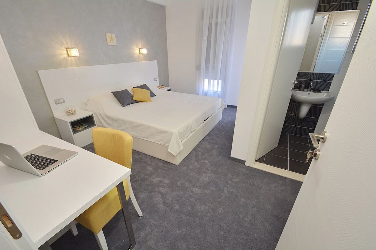 A hotel room with a bed, a desk with a laptop, a chair, a bedside table, and a bathroom with a sink and mirror.
