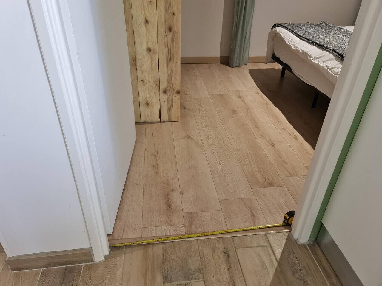 Ein Zimmer mit Holzboden, einem Bett mit weißer und grauer Bettdecke und einer Tür mit weißem Rahmen.
