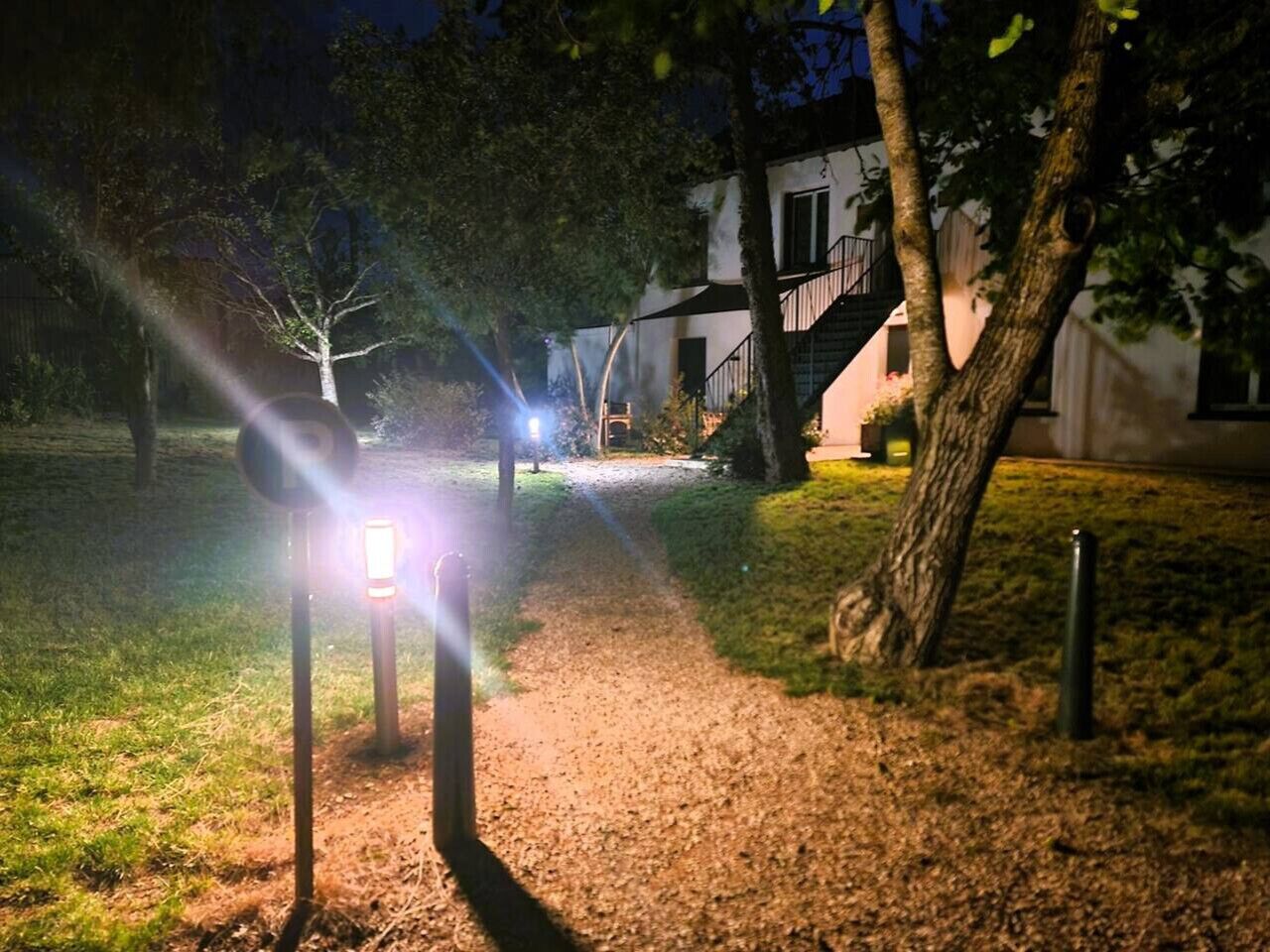 Ein beleuchteter Weg bei Nacht mit Leuchtpfosten, Bäumen und einem Gebäude mit einer Treppe im Hintergrund.