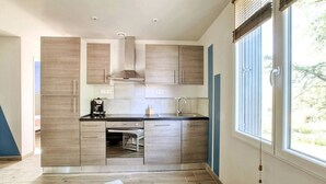 A modern kitchen with wooden cabinets, a built-in oven, and a window with blinds. - Cottage Duo Syrah Sauna Gaillac- LesCottagesduTarn (Brens)
