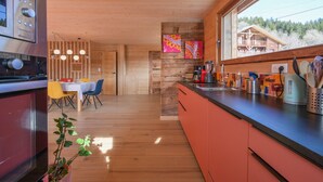 A modern kitchen with wooden cabinets, a dining area with a table and chairs, and a large window with a view of a building and trees.