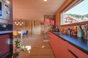 A modern kitchen with wooden cabinets, a dining area with a table and chairs, and a large window with a view of a building and trees. - Apartment in Chalet L'Albert with Mountain View, Balcony, and Private Terrace (Manigod)