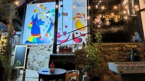An outdoor seating area with tables and chairs, a foosball table, and a mural with colorful abstract art. - Haku Hostel Cusco (Cusco)
