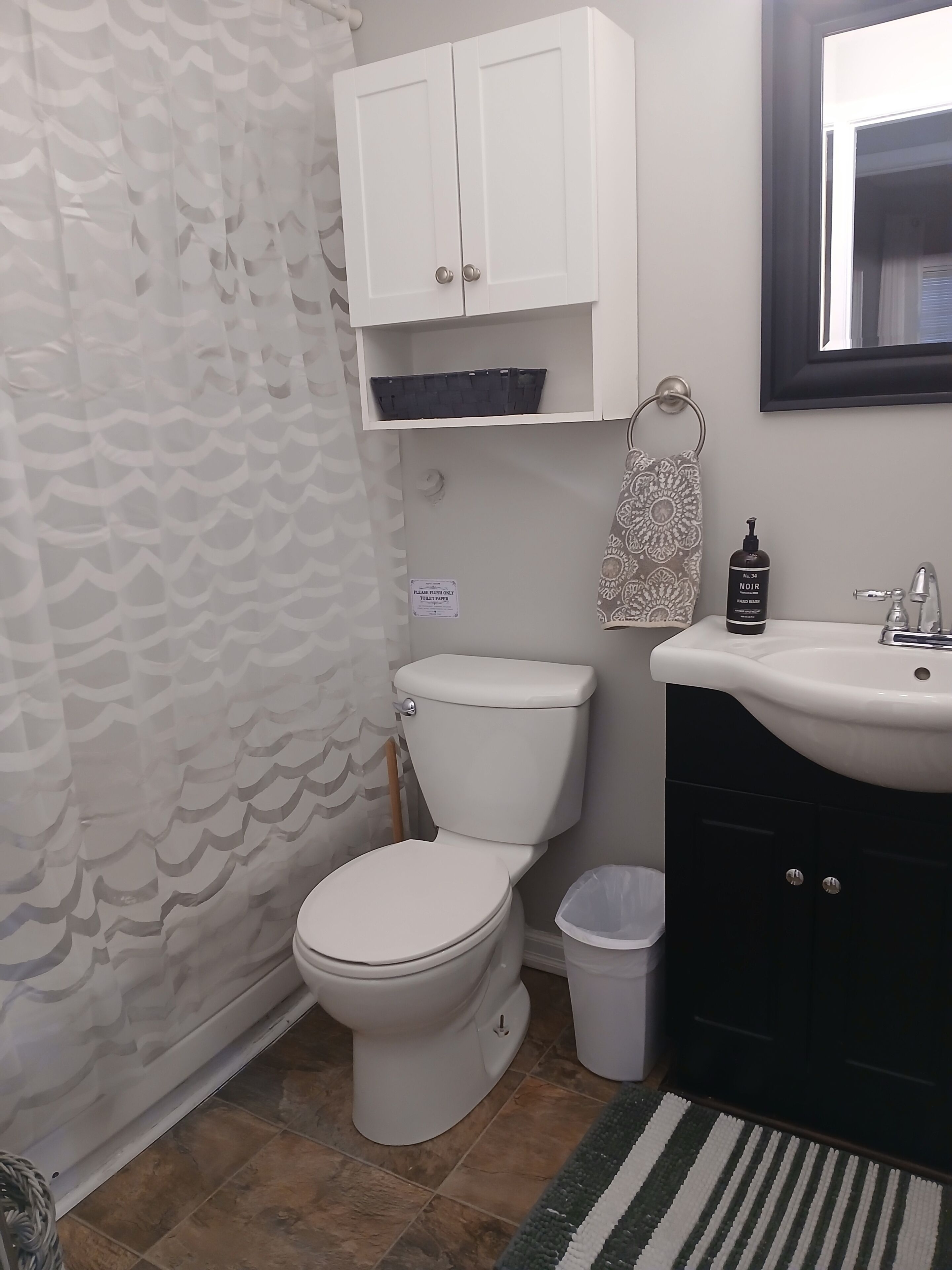 A bathroom with a toilet, a sink, a trash bin, a cabinet, and a patterned shower curtain.