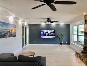 A spacious living area with a large flat-screen TV mounted on the wall, a ceiling fan, and a painting of a sunset on the wall. - The St. Pete Beach Home (Saint Petersburg)