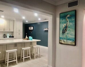 A kitchen with white cabinets, a bar area with stools, and a painting of a bird on the wall. - The St. Pete Beach Home (Saint Petersburg)