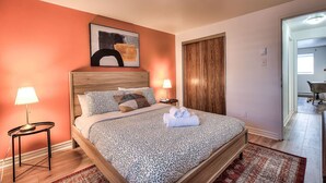 Une chambre à coucher avec un lit, deux lampes de chevet, une tête de lit en bois, un tableau abstrait encadré, une porte en bois et une vue sur une autre pièce.