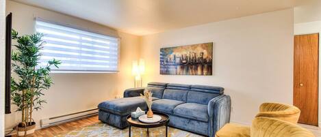 A living room with a blue sofa, yellow armchairs, a round coffee table, a cityscape painting, a potted plant, and a window with blinds.