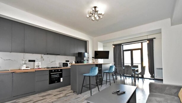 Een moderne keuken met grijze kastjes, een houten aanrechtblad en een inbouwoven. Er is een zithoek met een grijze bank, een salontafel en blauwe barkrukken. Een flatscreen televisie hangt aan de muur, en de kamer heeft grote ramen met gordijnen.