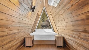 Ein raumförmiges, dreieckiges Holzgebäude mit einem Bett, zwei Nachttischen und einem Fenster mit Blick ins Freie.