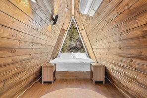 Ein raumförmiges, dreieckiges Holzgebäude mit einem Bett, zwei Nachttischen und einem Fenster mit Blick ins Freie.
